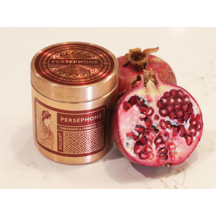 A bronze-colored candle vessel labeled 'PERSEPHONE' with an image of a woman, accompanied by a halved pomegranate revealing the seeds inside.