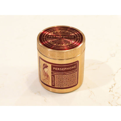 A bronze-colored candle vessel labeled 'PERSEPHONE' with an image of a woman scented with Pomegranate & Forbidden Love